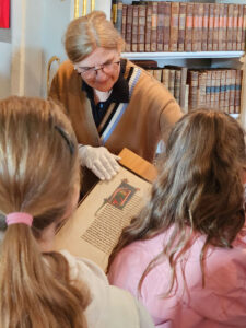Eine Archivmitarbeiterin zeigt einer Kindergruppe eine historische, reich verzierte Handschrift aus dem Mittelalter in einer Bibliothek mit alten Büchern.